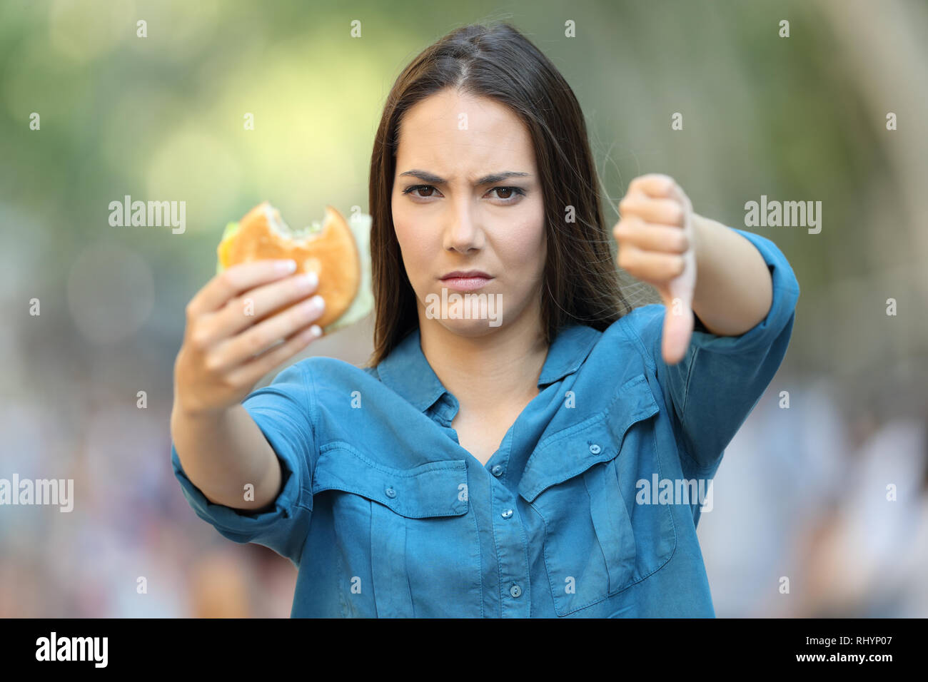 Front view of annoyed woman holding a burger with thumb down in the street Stock Photo