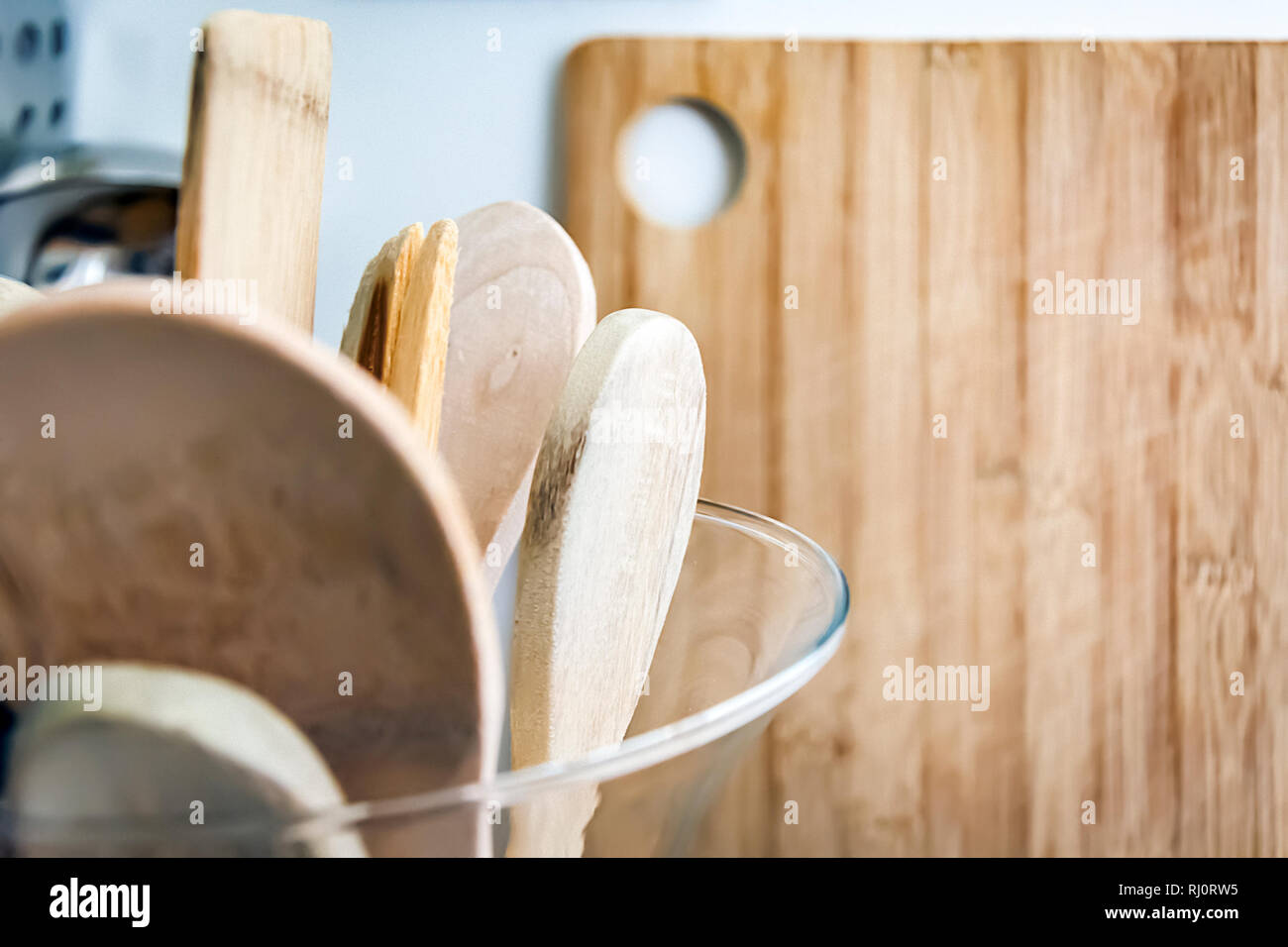 Wooden kitchen utensils in a glass container with a wooden cutting board in the background. Kitchen interior shot. Home decor and cooking concept Stock Photo