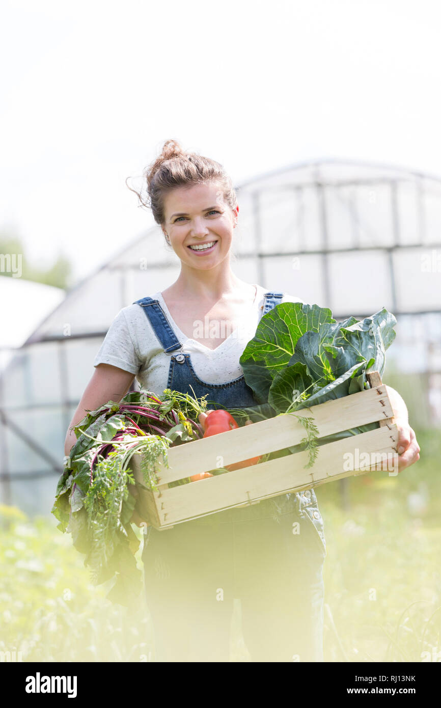 Portrait of smiling mid adult farmer holding vegetable crate at farm ...