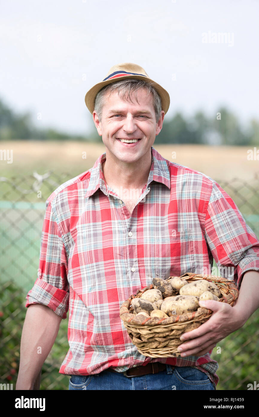 Portrait of smiling farmer carrying organic potatoes in basket at farm ...