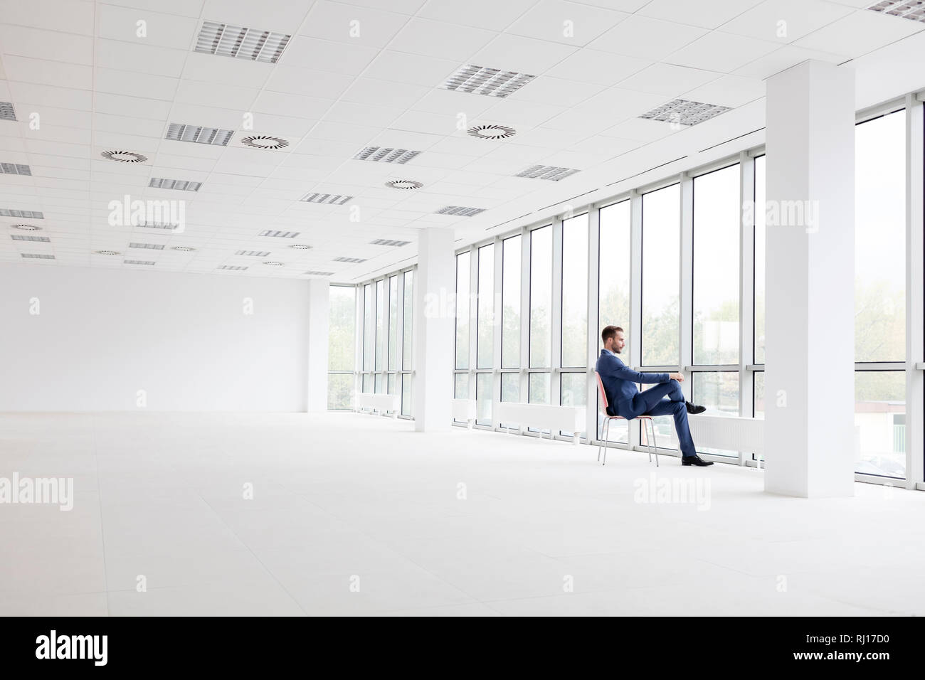 Full length of thoughtful young businessman sitting on chair while looking through window at new empty office Stock Photo