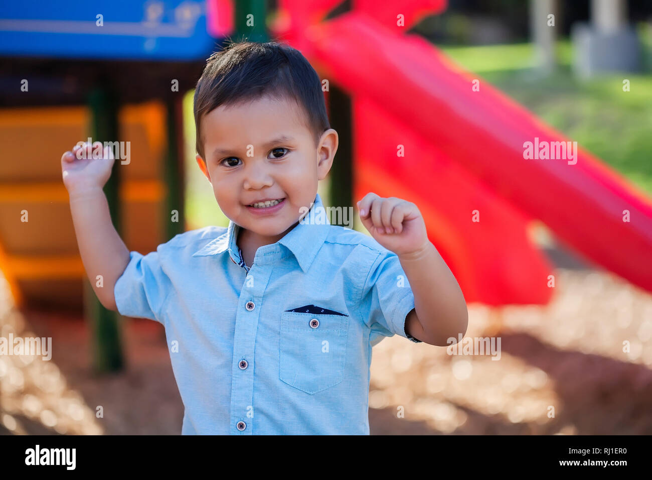 A toddler boy with arms raised up and a smile on his face, playing in a colorful kids playground. Stock Photo