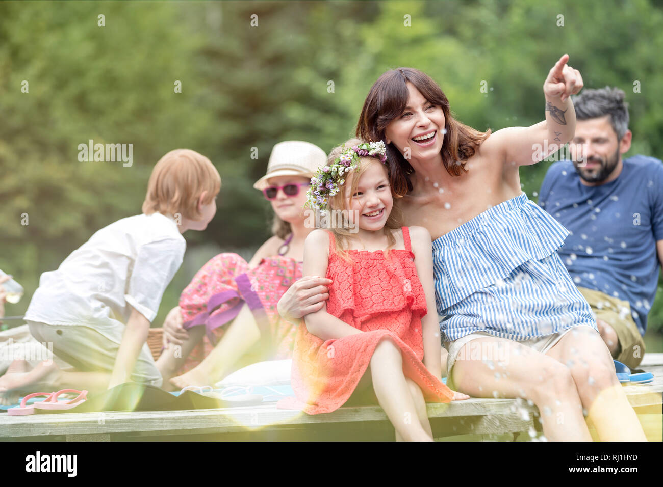 Smiling woman pointing to daughter while sitting on pier against family at lakeshore during summer Stock Photo