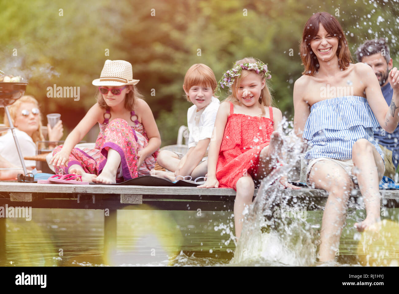 Smiling woman and daughter splashing water in lake while sitting on pier with family during summer picnic Stock Photo