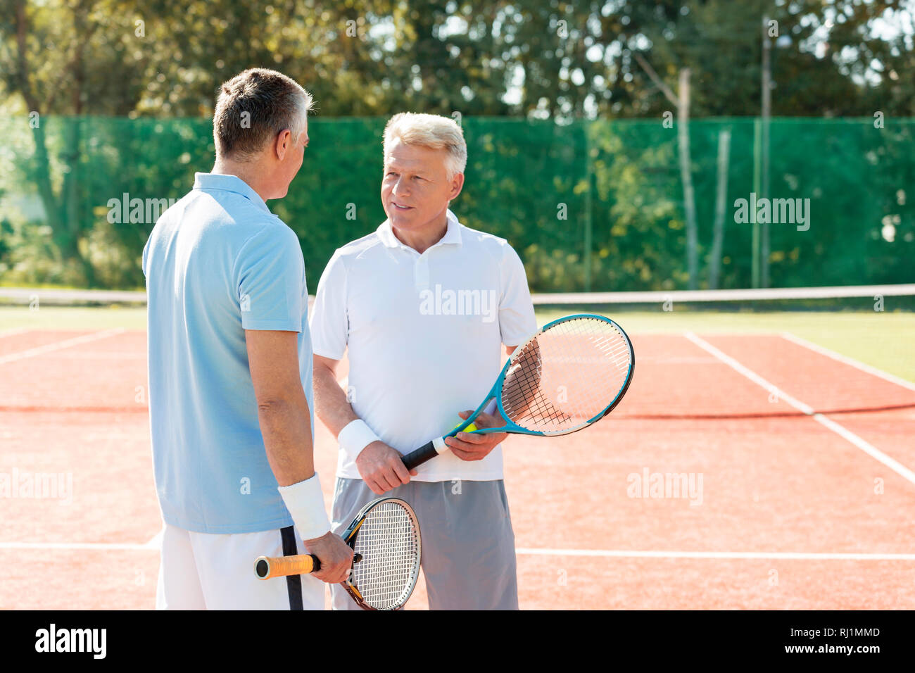 Mature men holding rackets while talking on tennis court during summer weekend Stock Photo