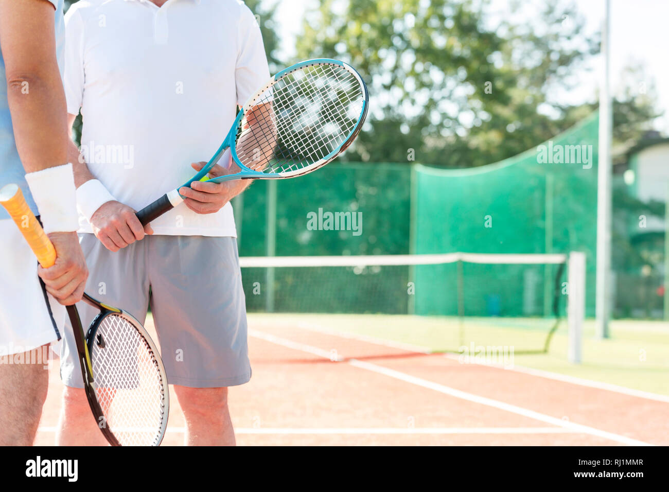 Midsection of mature men holding rackets while standing on tennis court during summer weekend Stock Photo