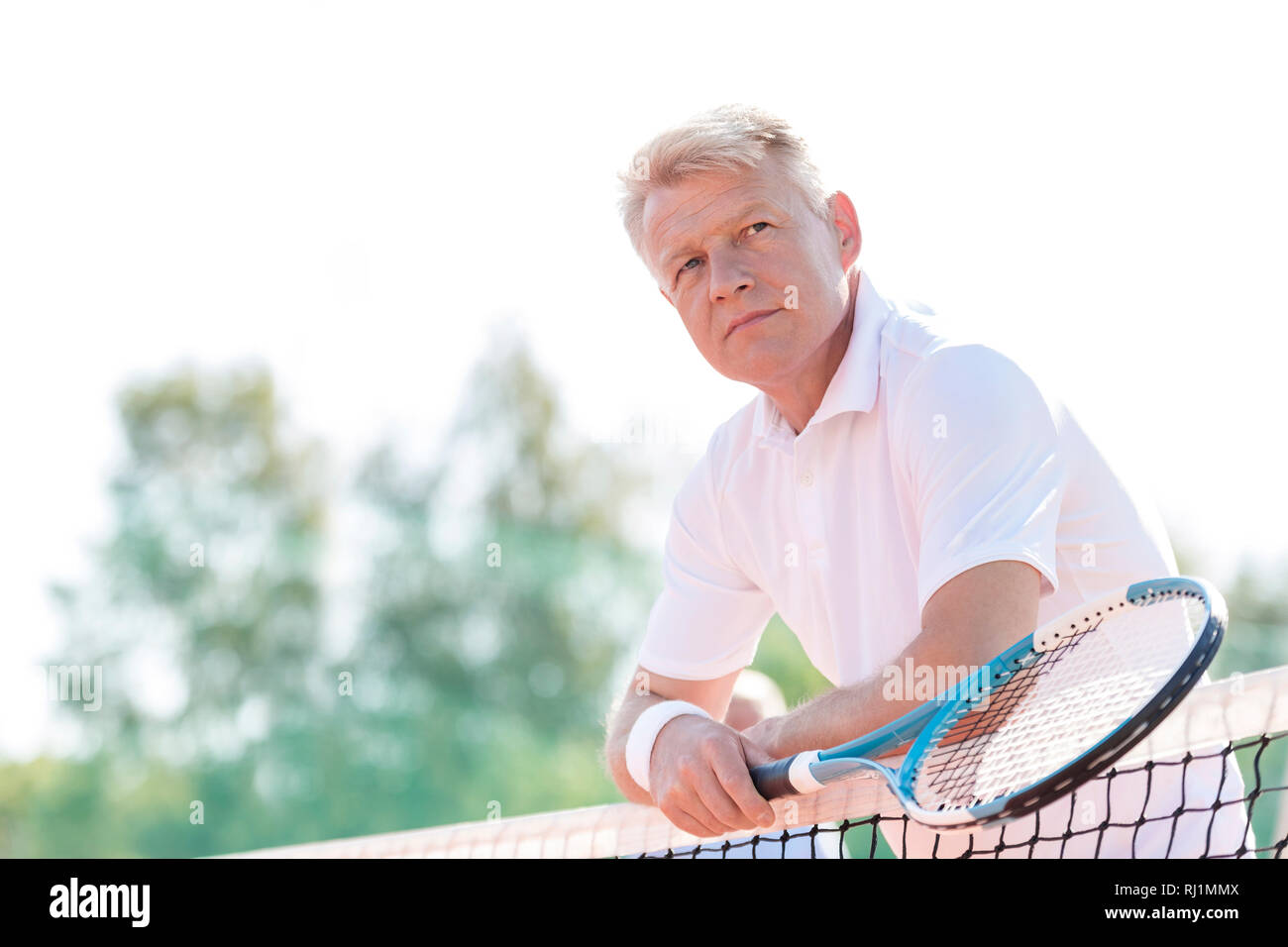 Thoughtful mature man holding tennis racket while leaning on net against clear sky Stock Photo
