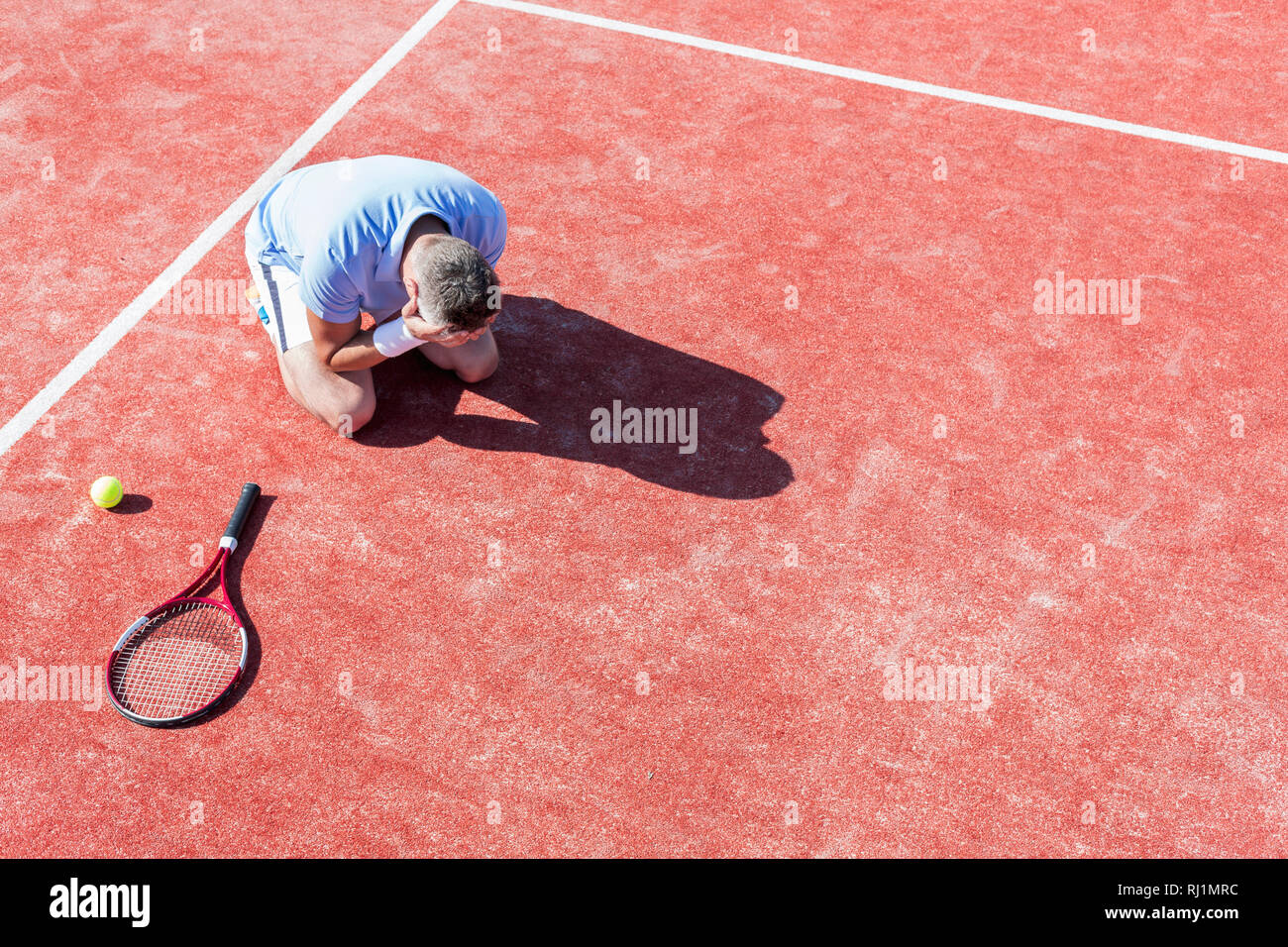 High angle view of disappointed mature man with head in hands while kneeling by tennis racket on red court during summer Stock Photo