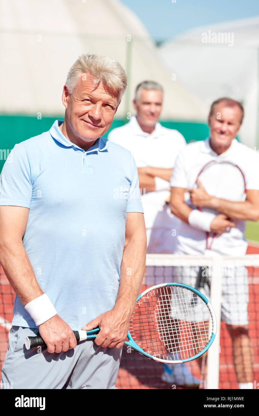 Portrait of confident mature man holding tennis racket while standing against friends on court during sunny day Stock Photo