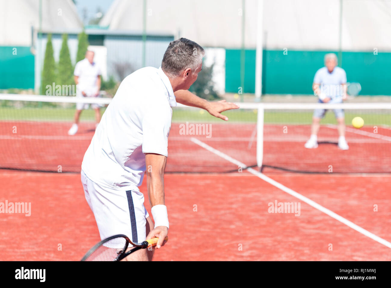 Man swinging racket while playing tennis doubles on red court during summer weekend Stock Photo