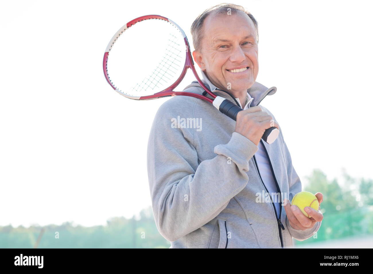 Portrait of smiling retired senior man standing with tennis racket and ball against clear sky on sunny day Stock Photo