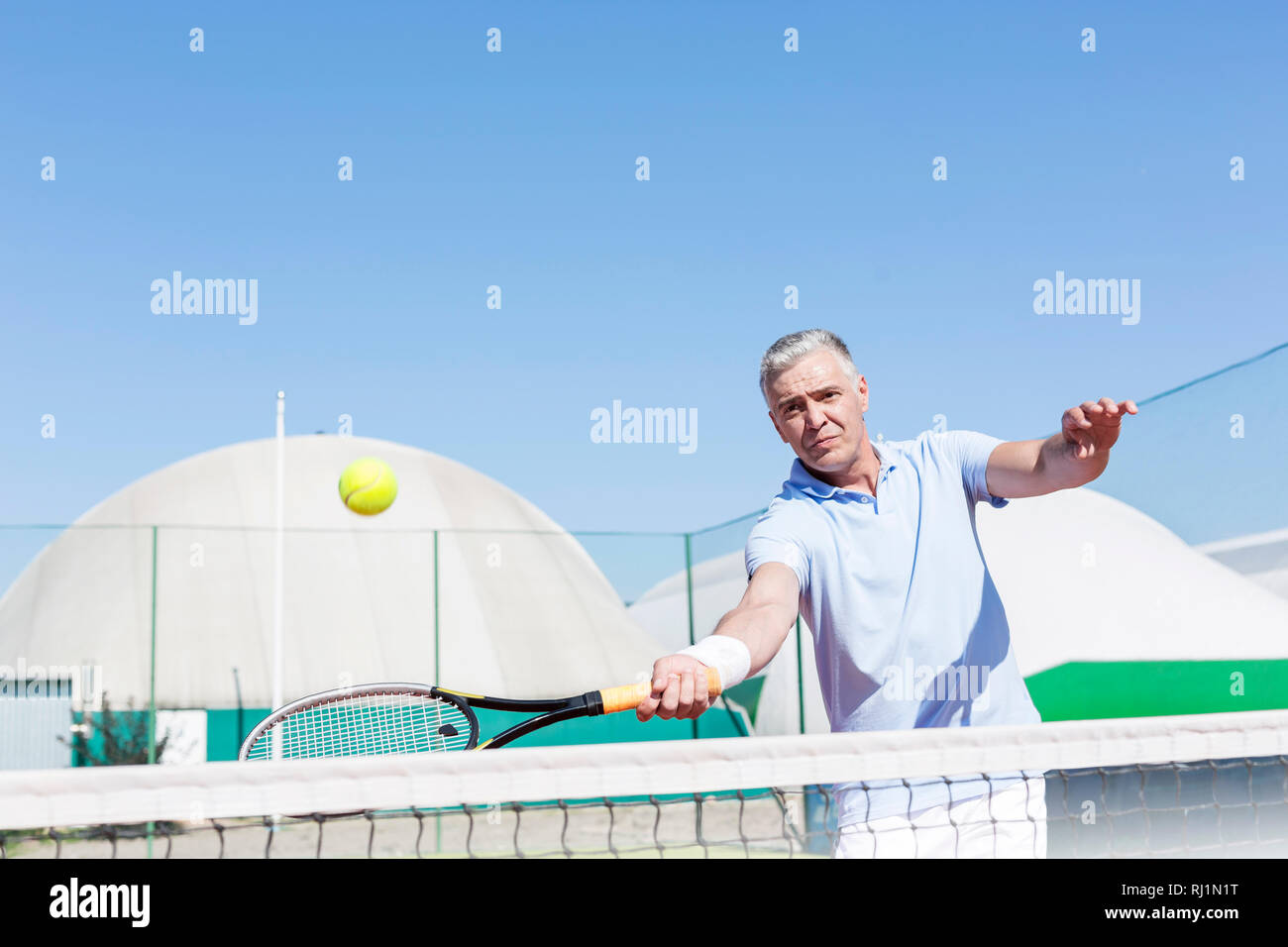 Confident mature man hitting tennis ball with racket on court against clear blue sky Stock Photo