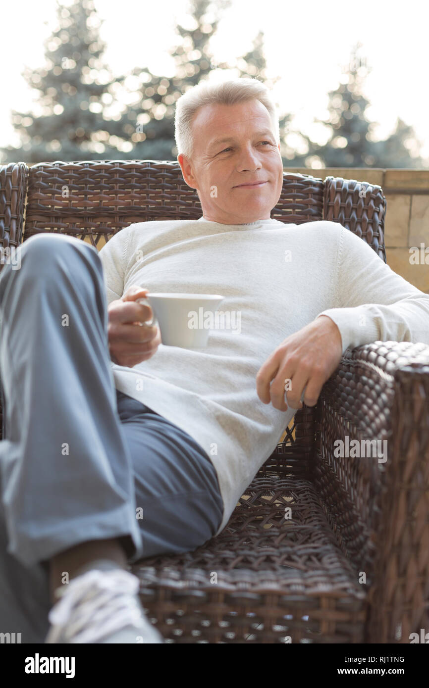 Smiling mature man holding drink while sitting on wicker chair at patio Stock Photo