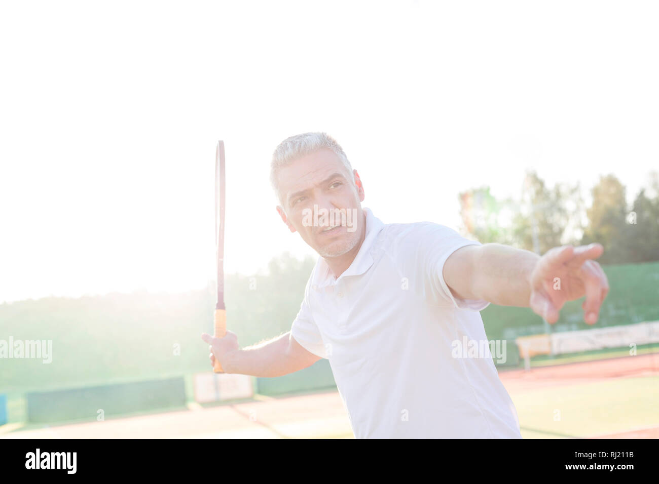 Back lit of mature man swinging tennis racket on court against clear sky Stock Photo
