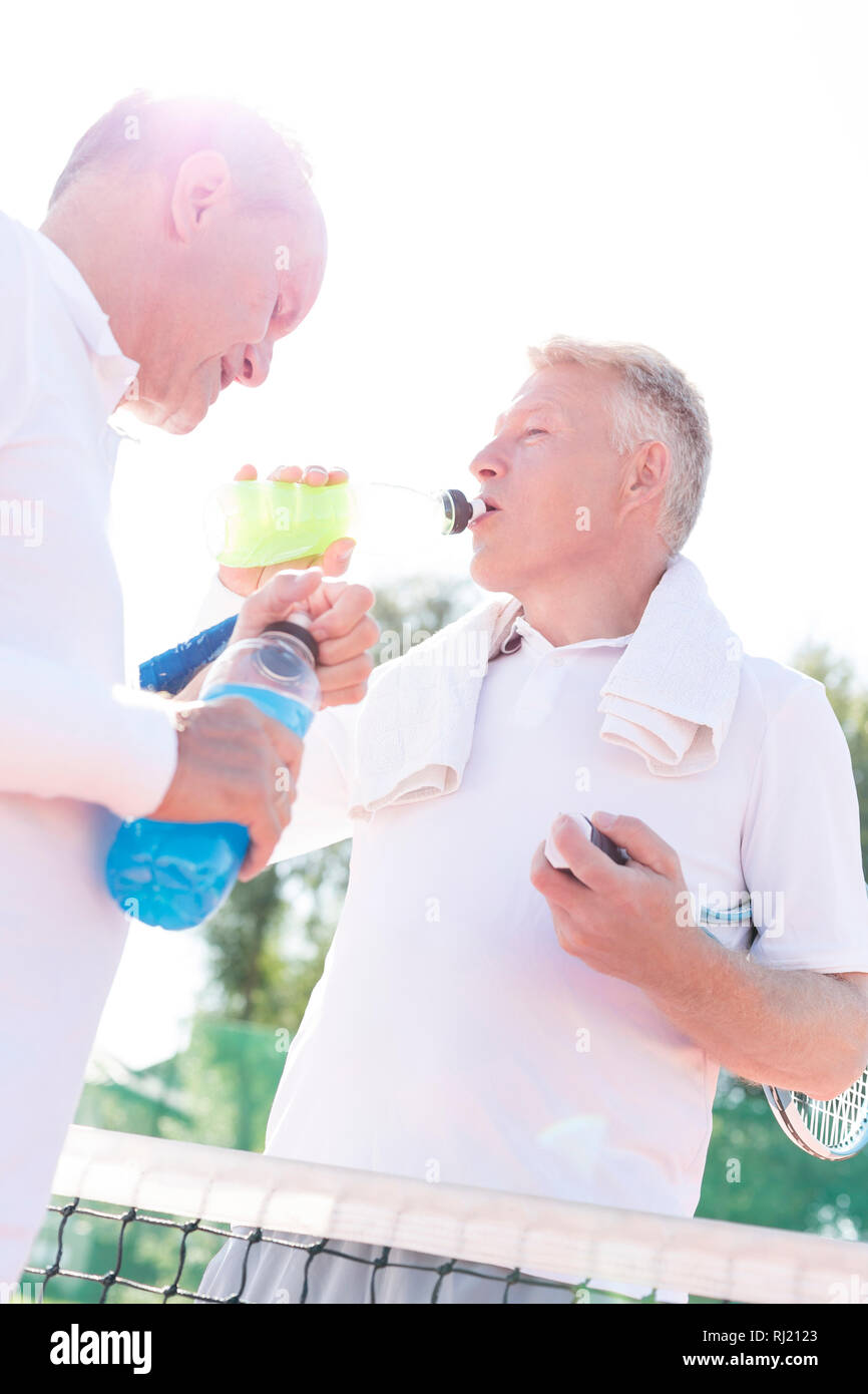 Low angle view of thirsty men drinking while standing by senior friend on tennis court against clear sky Stock Photo