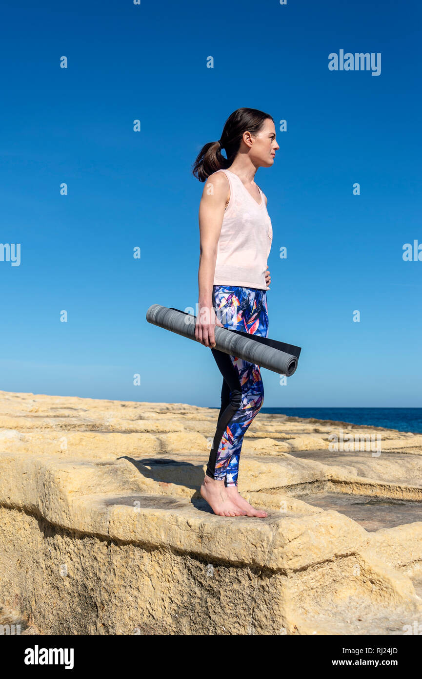 sporty woman carrying a rolled up yoga mat standing on rocks looking