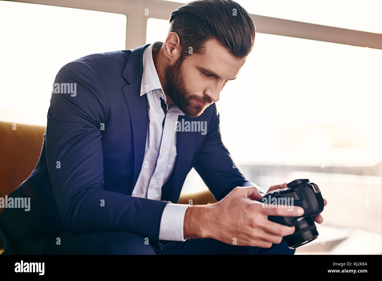 Close-up portrait of handsome young businessman in fashion suit who is looking at pictures on camera while sitting on sofa at office Stock Photo