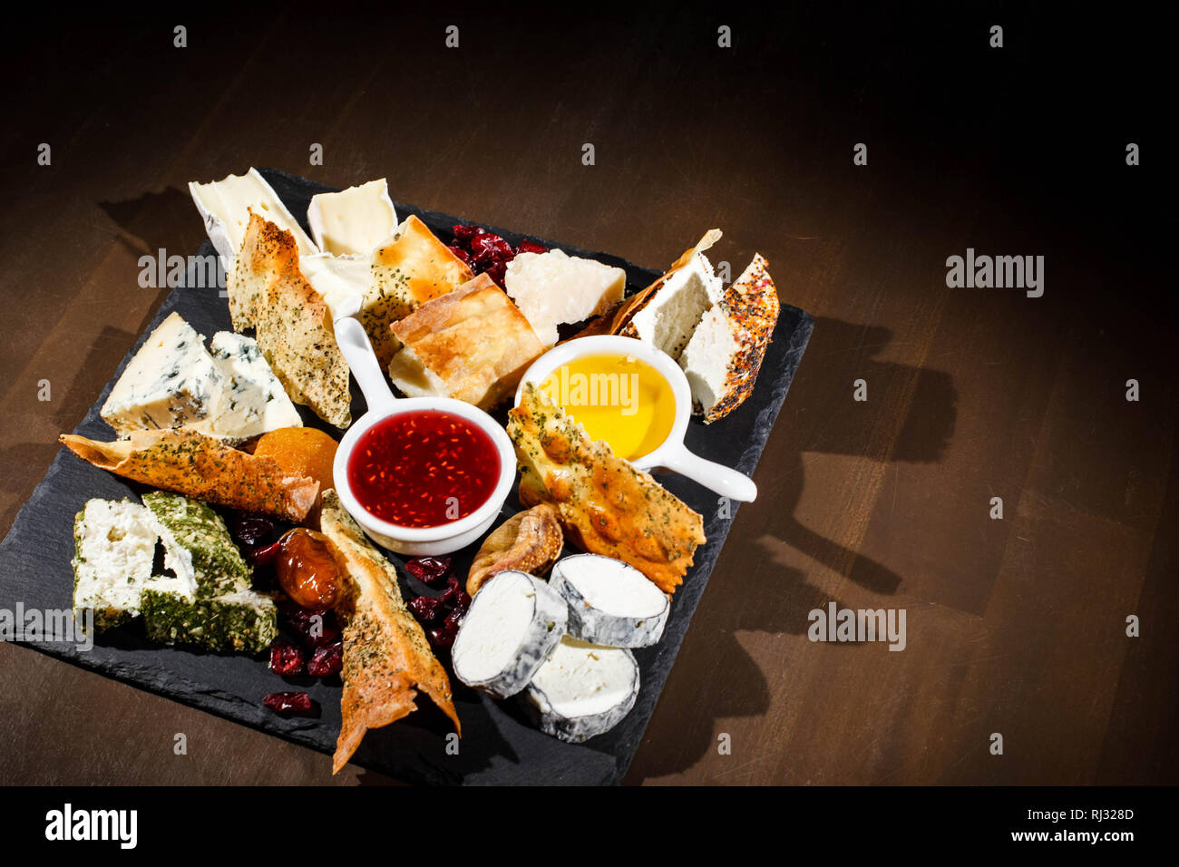 Pieces of camembert,  blue cheese, cheddar lie on black plate with sauces in white bowls Stock Photo