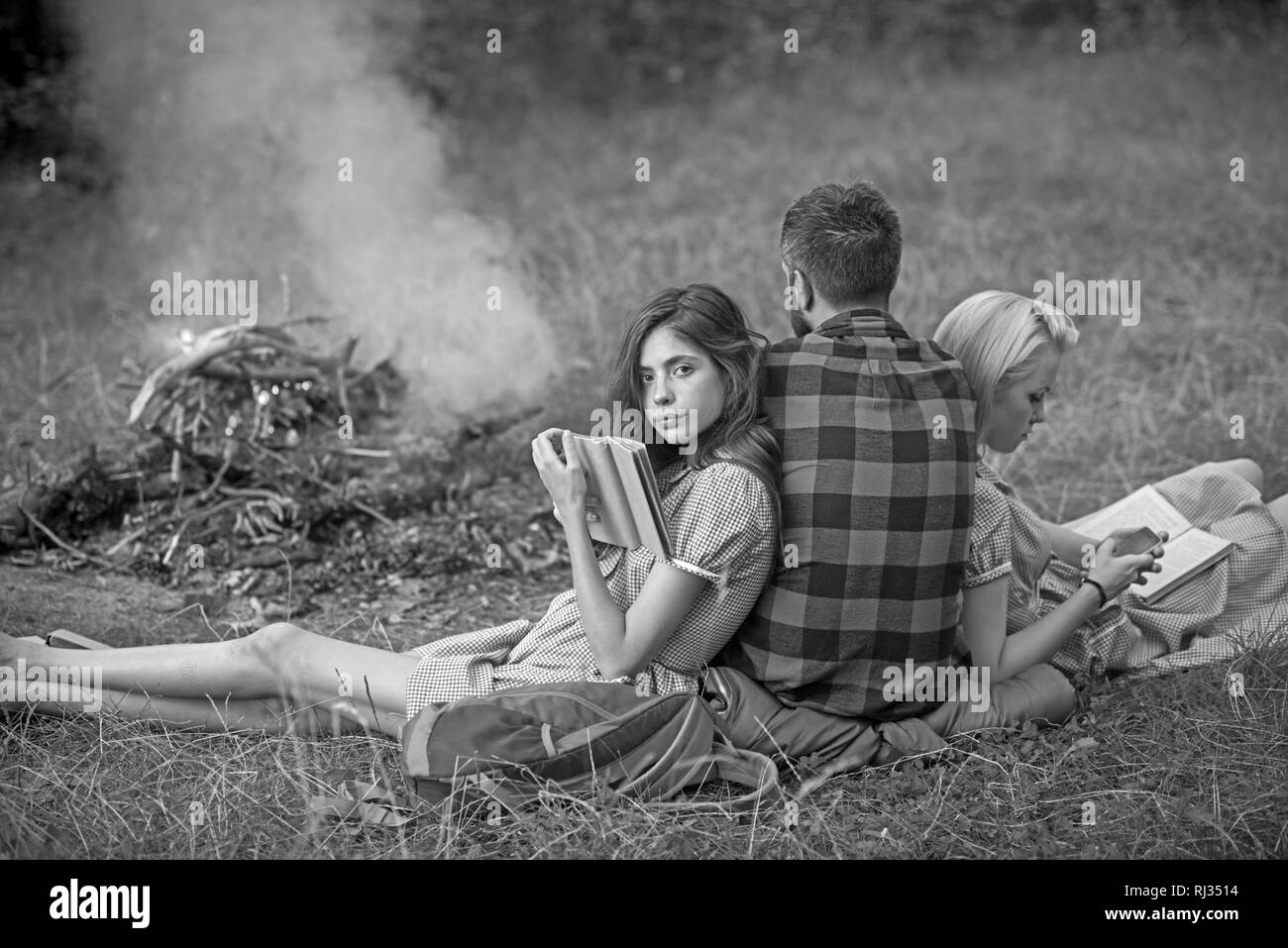 Camping in wilderness. Turn back guy looking at fire while two beautiful girls read book, sustainable education concept Stock Photo