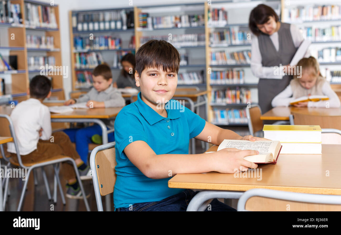 Portrait of smiling intelligent preteen boy reading in school library on background with other students and teacher Stock Photo
