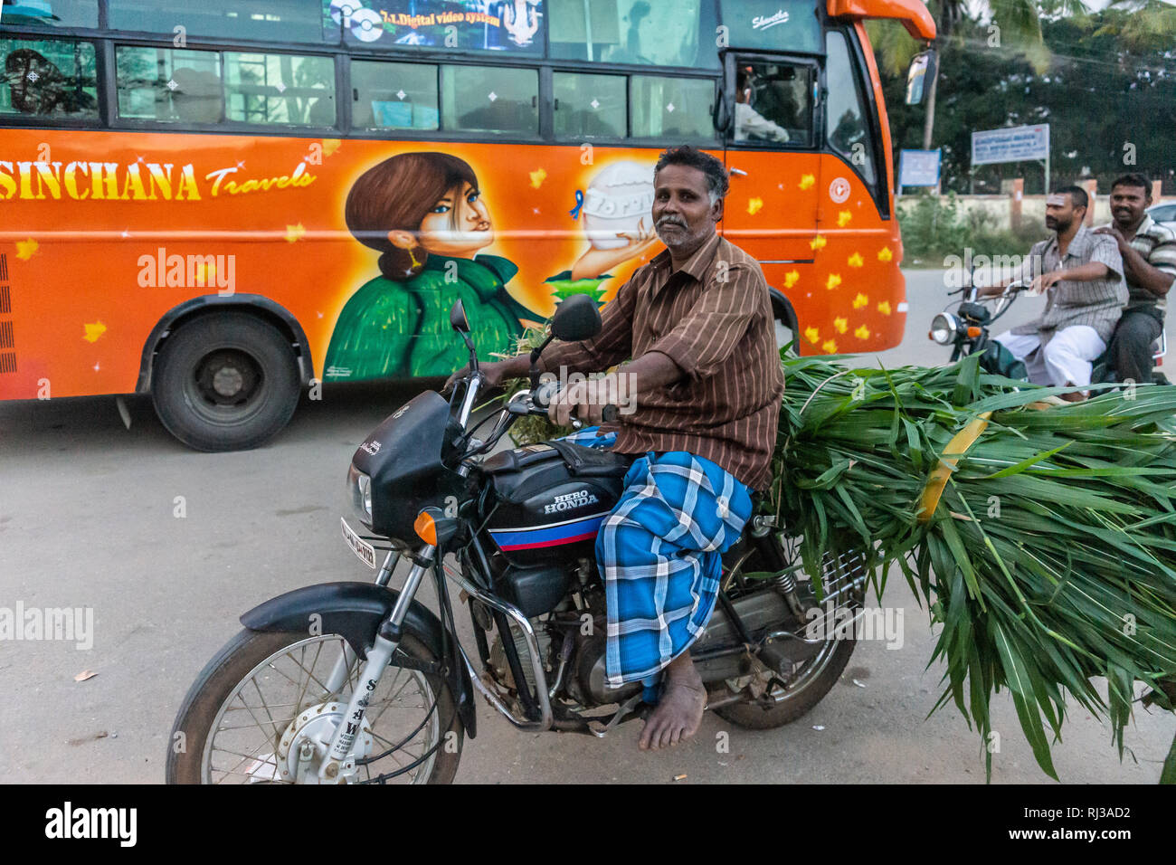 Halebidu, Karnataka, India - November 2, 2013: Street scene in town with orange public bus and motorcycles, one overloaded with green sugar cane stalk Stock Photo