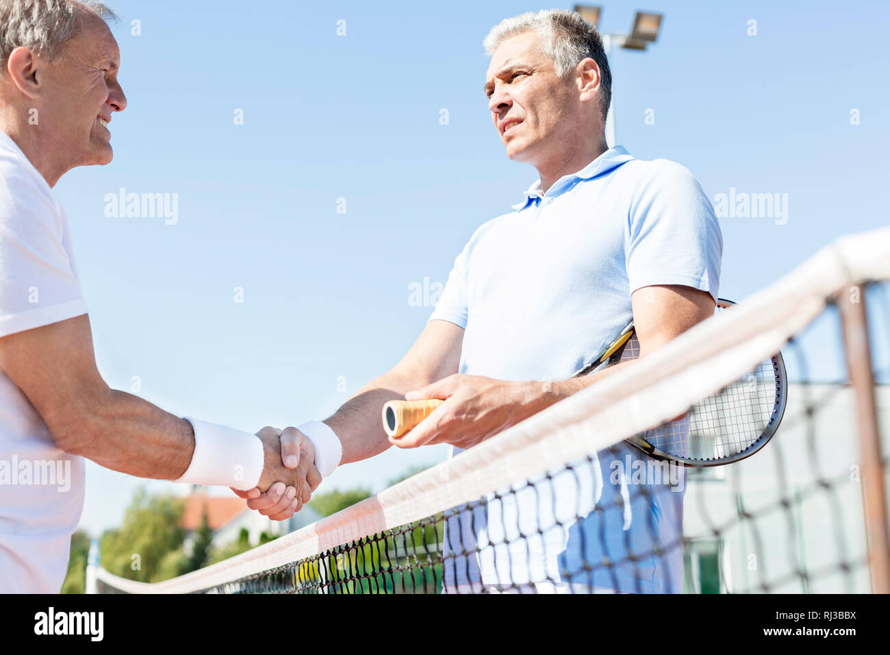 Men shaking hands while standing by tennis net against clear sky on sunny day Stock Photo