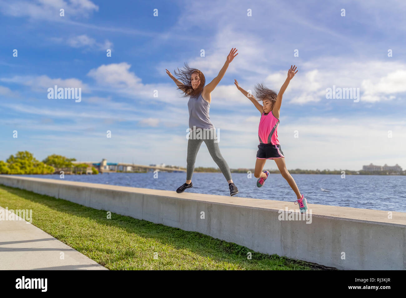 Mother and daughter jump together from a seawall on a bright sunny day. Stock Photo