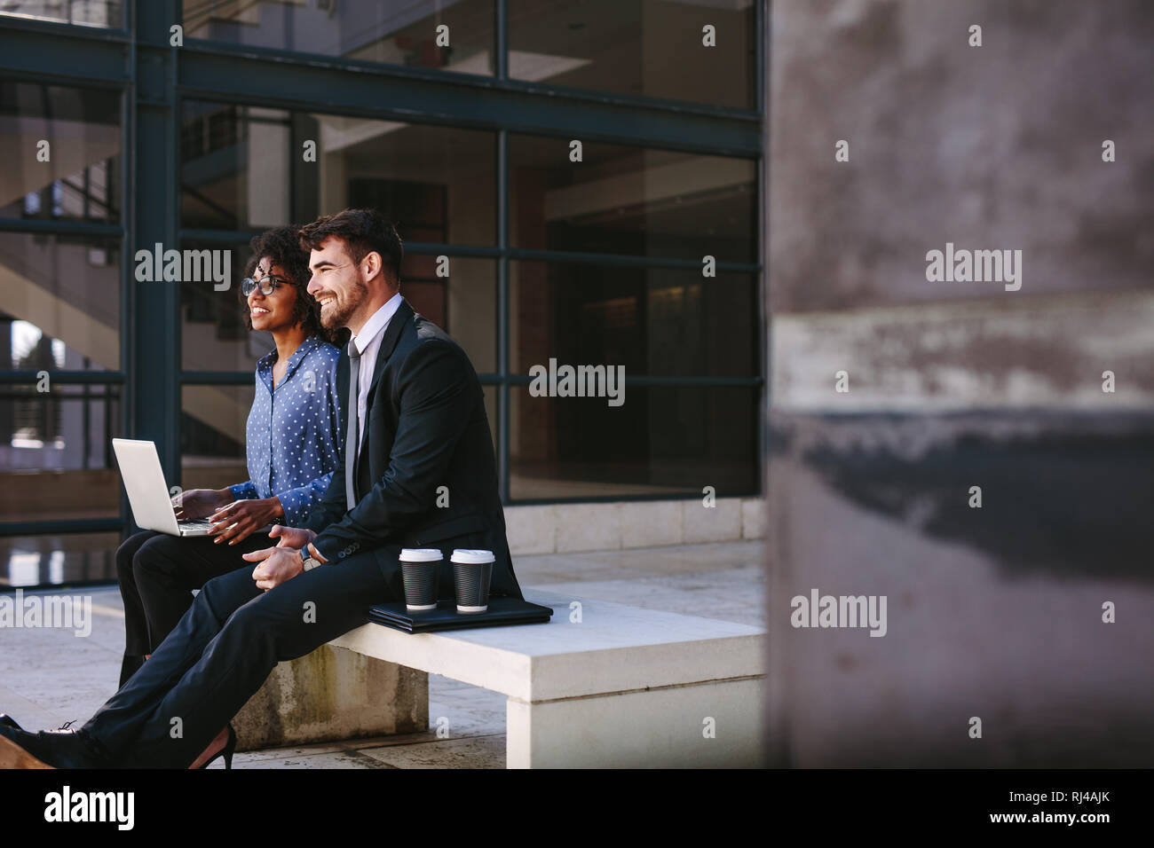 business colleagues sitting at bench at office lobby with laptop and looking away smiling. businessman and woman relaxing at office hallway with coffe Stock Photo