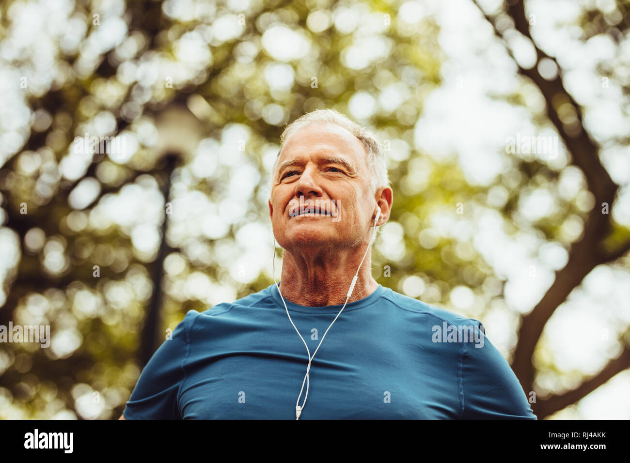 Low angle shot of a senior man standing under a tree listening to music during workout. Close up of a senior fitness person doing workout outdoors. Stock Photo