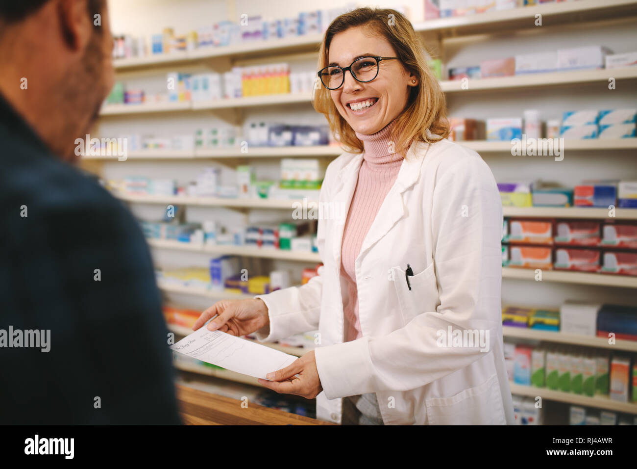 Smiling female pharmacist with prescription assisting a customer standing at the counter. Helpful female pharmacist dealing with a male customer stand Stock Photo