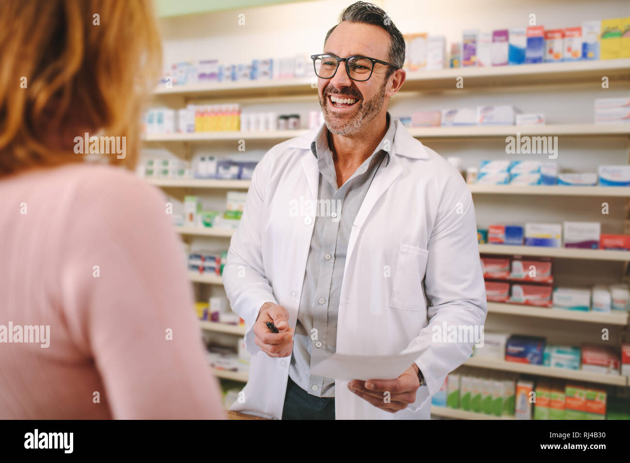 Helpful male pharmacist dealing with a woman customer standing at desk in the pharmacy. Smiling male pharmacist with prescription assisting a customer Stock Photo