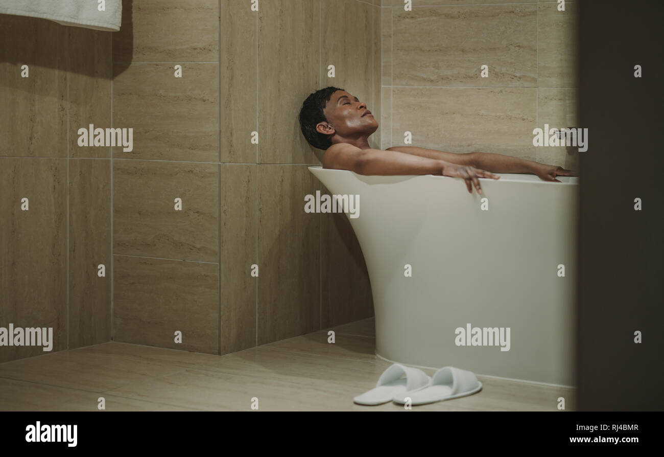 Mature african woman lying in bathtub with her eyes closed. Female relaxing in bathtub. Stock Photo