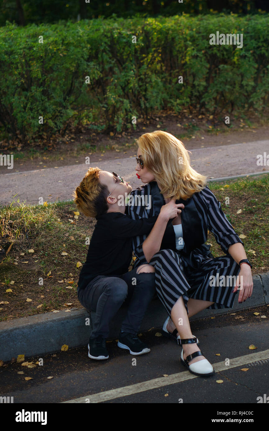 Young attractive mother and son are sitting on curb in city park. Boy