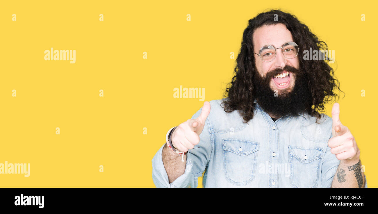 Young hipster man with long hair and beard wearing glasses pointing fingers to camera with happy and funny face. Good energy and vibes. Stock Photo