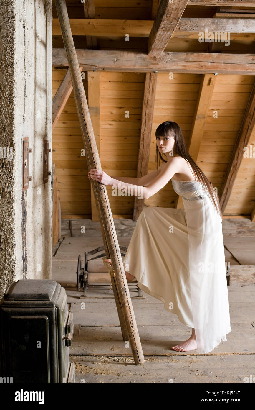 Frau steht mit Abendkleid vor einer Leiter auf dem Dachboden Stock Photo