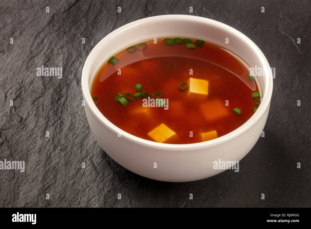 A bowl of miso soup with tofu and scallions on a black background with
