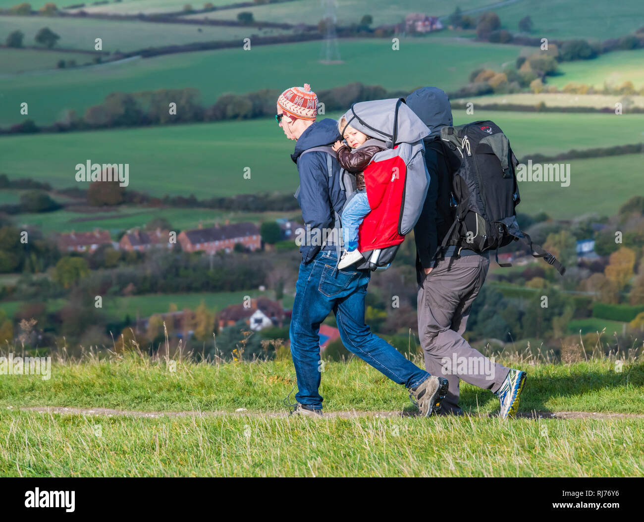 Couple of people walking in the countryside carrying a child in a child carrier backpack wearing hats & coats on a cold day in Autumn, West Sussex, UK Stock Photo