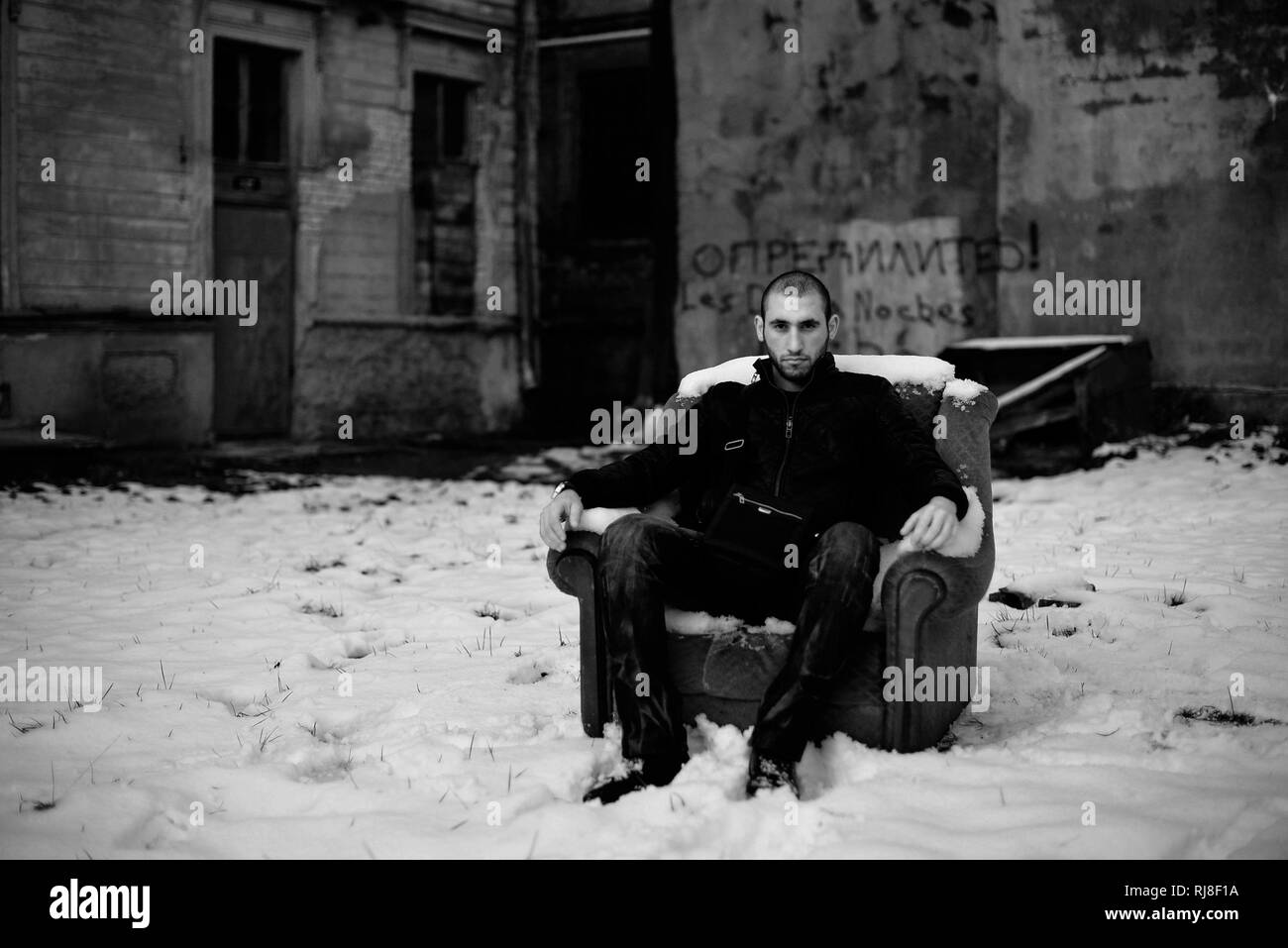 Black and white portrait of a bold guy sitting on abandoned chair on the street by an old house in ghetto. Winter. Snow. Stock Photo