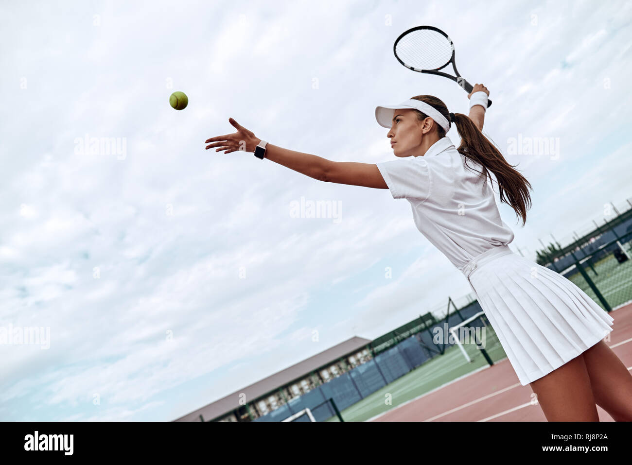 Attractive sportswoman in white sportswear with a racket in her hand throws the ball into the air on the tennis court Stock Photo