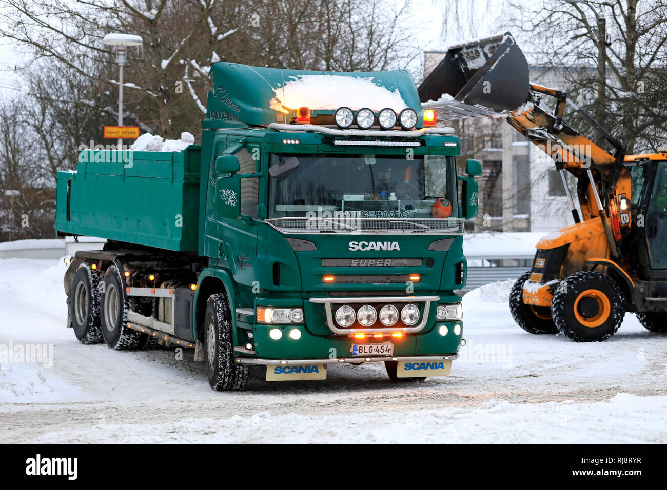 Salo, Finland - February 2, 2019: JCB loader loads snow onto green Scania P420 dump truck trailer to be transported to a snow dumping area. Stock Photo