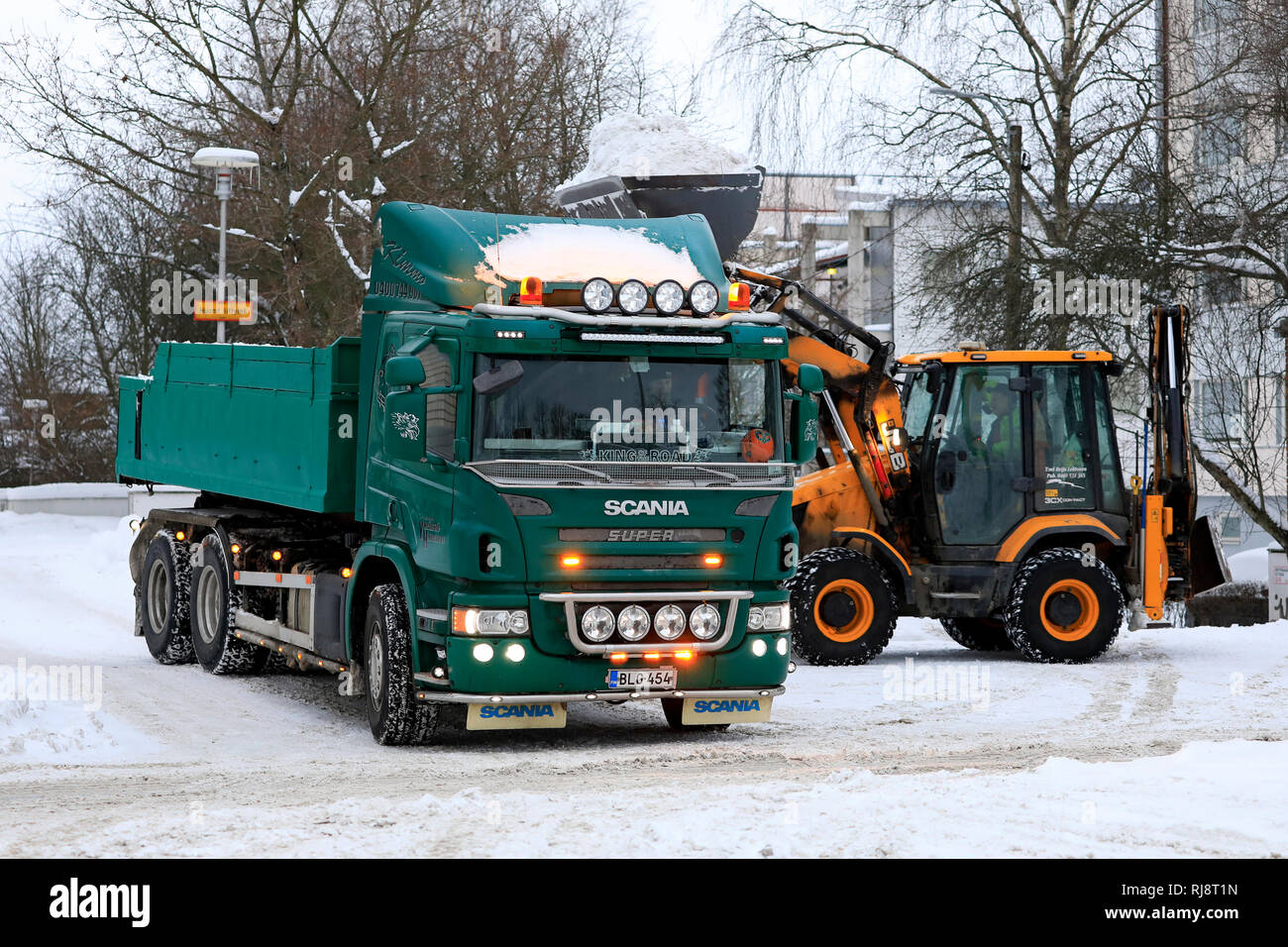 Salo, Finland - February 2, 2019: JCB loader loads snow onto green Scania P420 dump truck trailer to be transported to a snow dumping area. Stock Photo