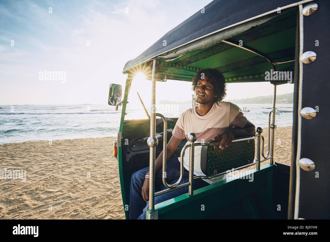 Young tuk tuk driver waiting for passenger against sand beach and sea in Sri Lanka. Stock Photo