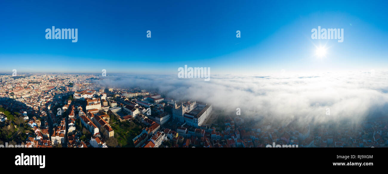 Aerial; panoramic drone view of foggy Lisbon morning; impending clouds over the ancient ginger roofs; blue skyline and bright sun in portuguese histor Stock Photo