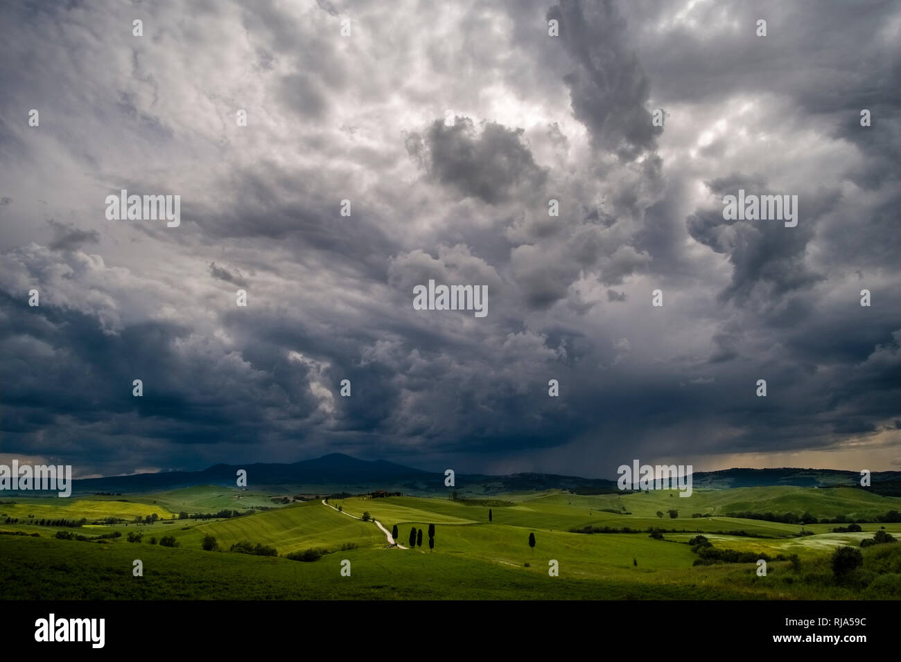 Panoramic aerial view on a typical hilly Tuscan countryside in Val d’Orcia with fields, trees, cypresses and a farmhouse, dark thunderstorm clouds app Stock Photo