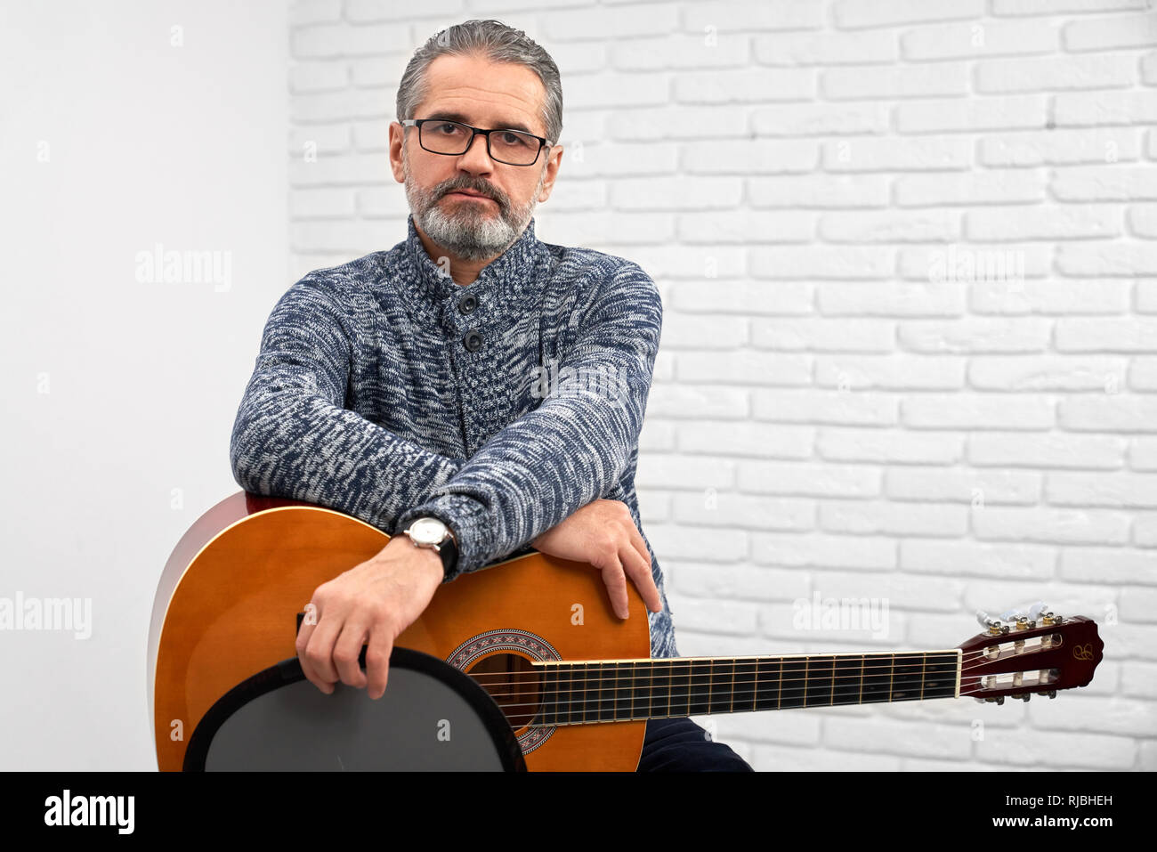 Handsome mature man sitting and holding acoustic guitar on knees. Bearded musician wearing glasses and watch. Man looking at camera and posing in studio with musical instrument. Stock Photo