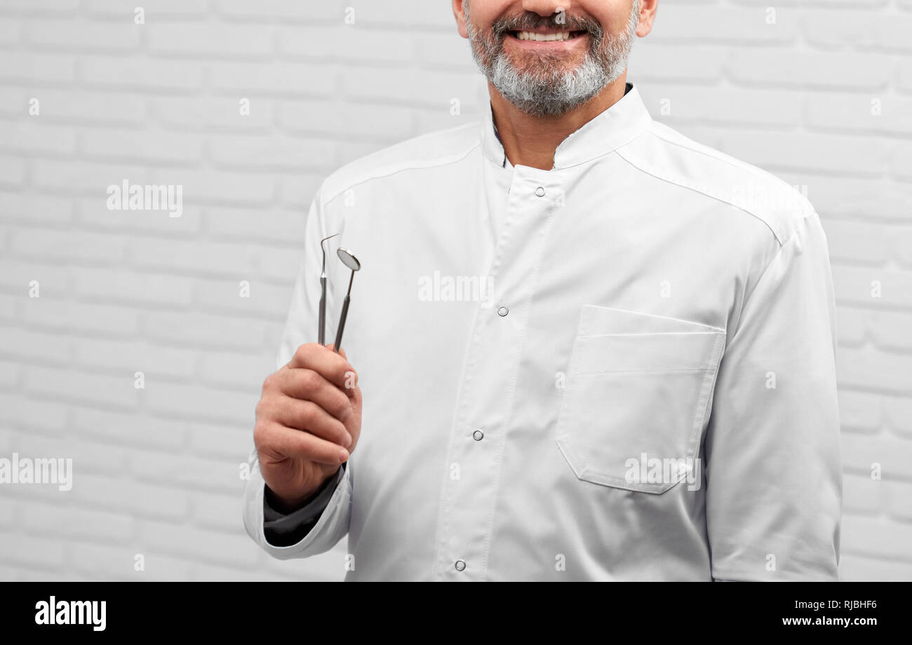Cropped potrait of happy dentist wearing in white medical uniform. Bearded doctor holding restoration instruments. Professional specialist smiling, posing with tools in hands. Stock Photo