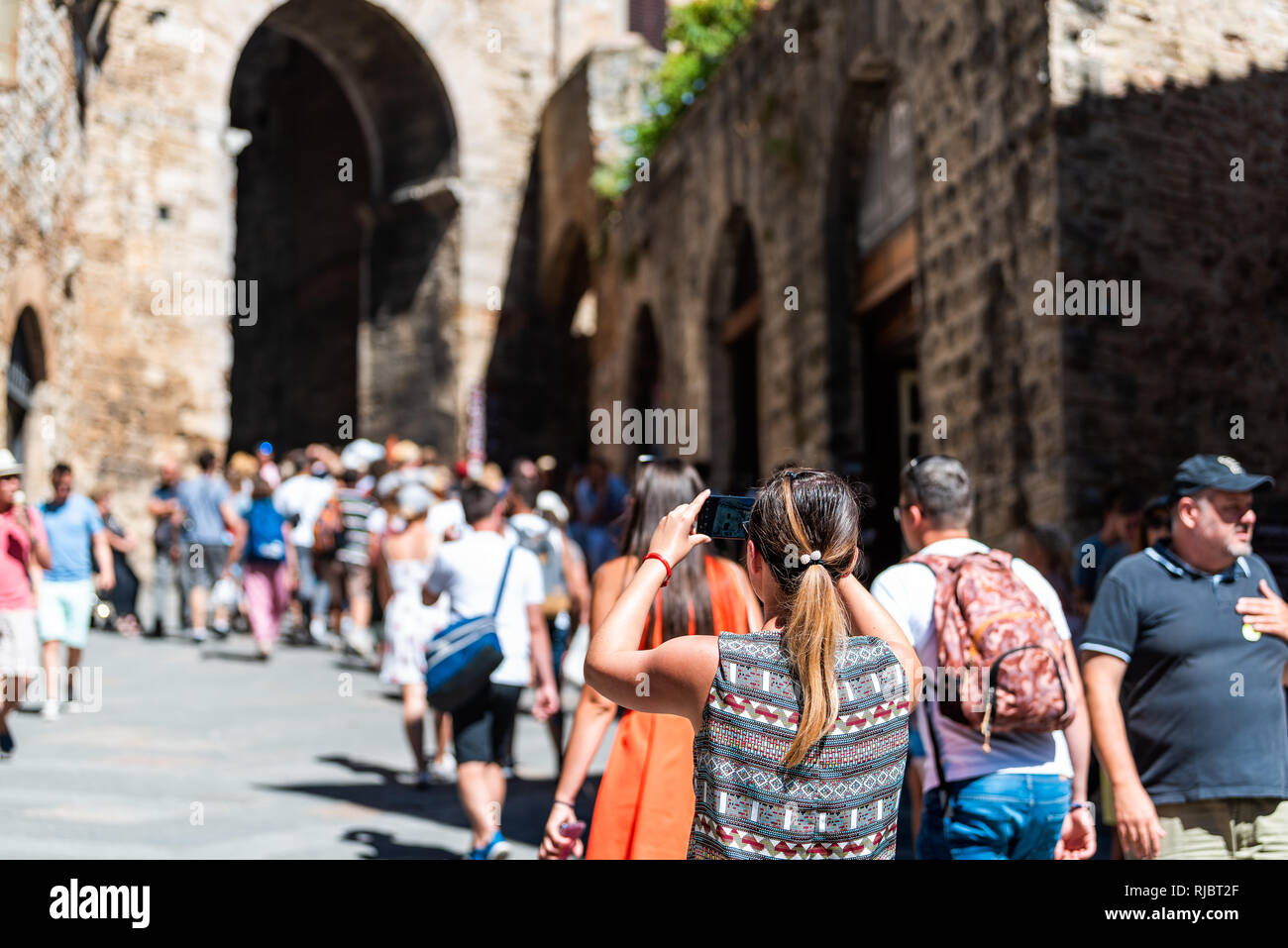 San Gimignano, Italy - August 27, 2018: Back of many crowd of people tour group walking exploring arch of small historic medieval town village in Tusc Stock Photo