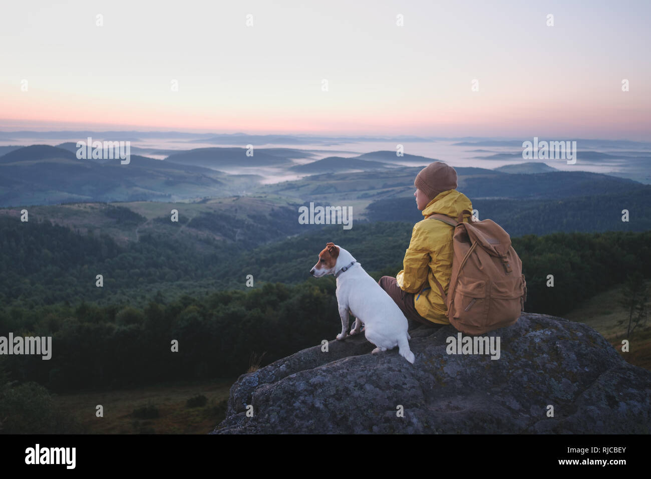 Alone tourist sitting on the edge of the cliff with dog against the backdrop of an incredible mountain landscape. Sunny day and blue sky Stock Photo