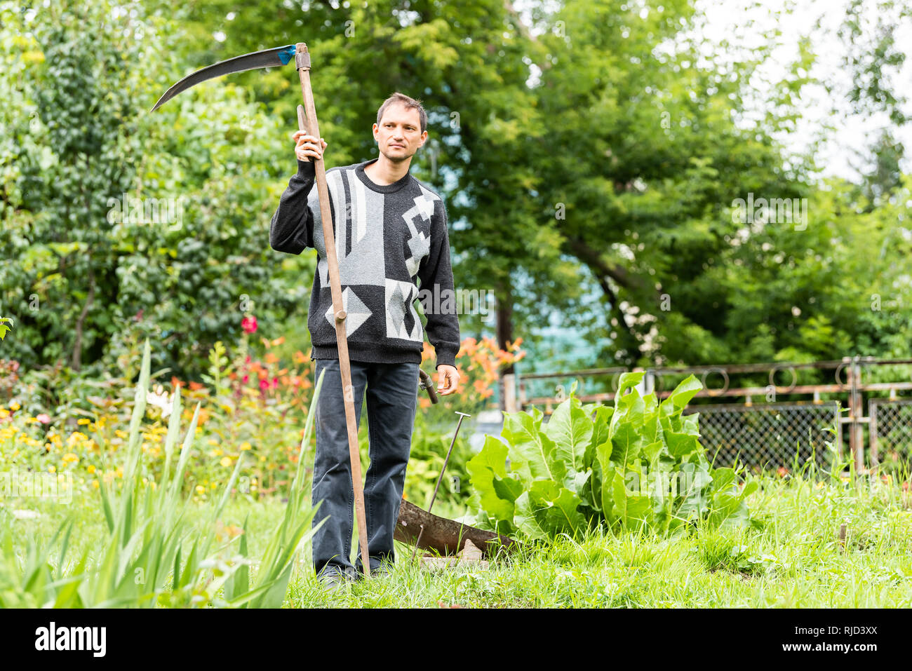 Back of young man farmer in garden standing with sickle scythe rake tool in green summer in Ukraine by well faucet Stock Photo