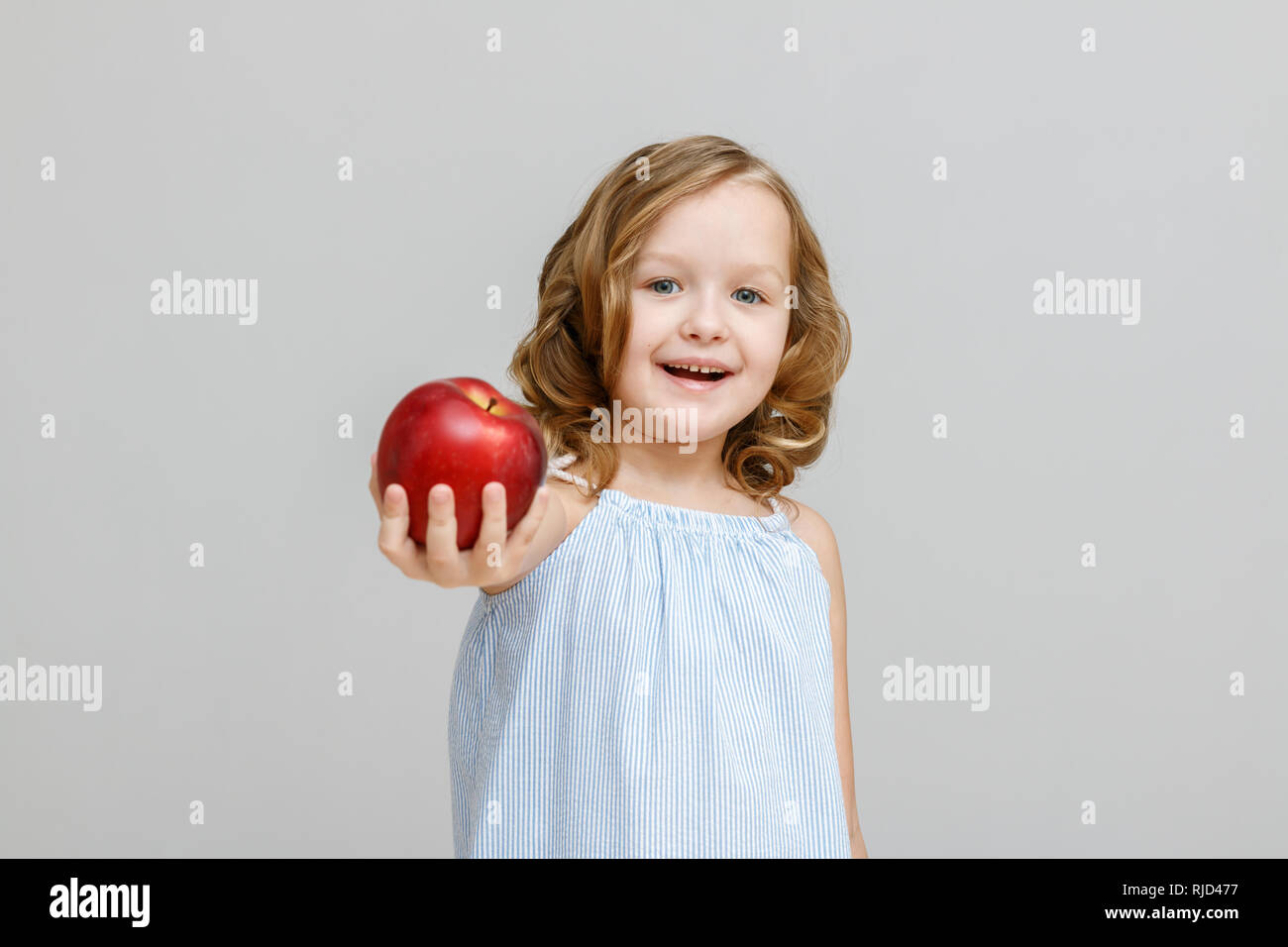 Portrait of a happy smiling little blonde girl on a gray background. A child holds out a red apple Stock Photo